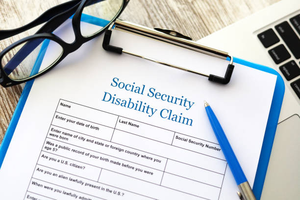 A blue clipboard contains an application for Social Security Disability Benefits. A blue pen and a pair of reading glasses sit on top of the clipboard.